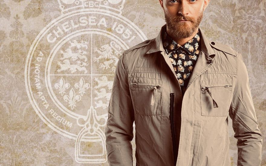 A bearded man with curly hair wearing a light khaki Chelsea Harbour jacket layered over a floral button-up shirt. He stands confidently in front of a vintage-style Chelsea Boot Co. crest.
