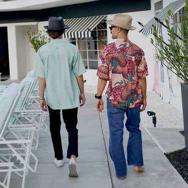 Two men walking outdoors in Da Vinci California camp shirts - one in a color-block style, the other in a Koi-print shirt and cowboy hat.